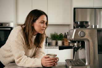 Femme examinant une machine à café expresso dans une cuisine moderne en marbre blanc, tenant un mug en céramique